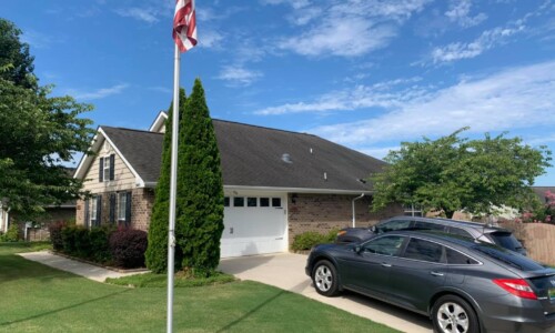 A brick suburban house with a two-car garage, two parked cars, and an American flag on a sunny day.