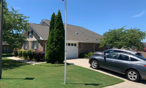 Single-story brick house with a front yard, driveway, two parked cars, and a flagpole under a clear blue sky.