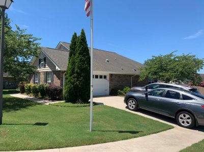 Single-story brick house with a front yard, driveway, two parked cars, and a flagpole under a clear blue sky.