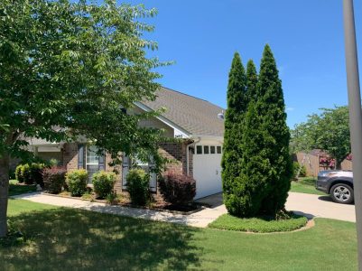 A single-story brick house with a two-car garage, manicured lawn, shrubs, and a tall tree on a sunny day.