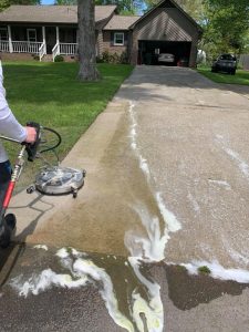A person pressure washes a concrete driveway, with a clear line between cleaned and uncleaned sections.