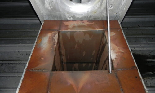 A close-up of a rusted metal ventilation duct opening with visible welds and a rope hanging beside it.