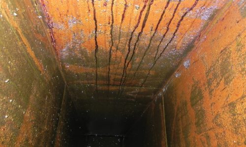 Rusty, rectangular metal duct or shaft interior with vertical streaks and a dark, grimy surface.