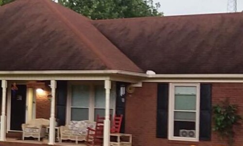 Single-story brick house with a covered porch, two red rocking chairs, and several plants in front.