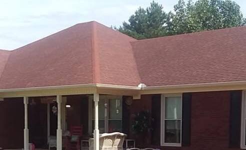 Single-story red brick house with a red roof, front porch, white columns, and outdoor furniture.