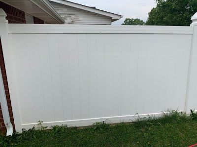 A white vinyl privacy fence panel with grass and weeds growing at the base, next to a brick house.