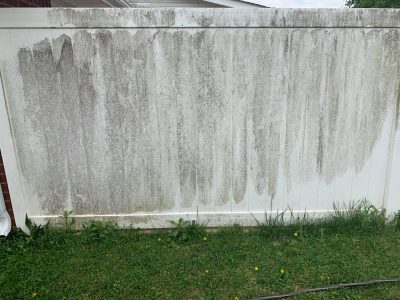 A white vinyl fence with heavy dirt and grime streaks, standing on green grass with some yellow dandelions.