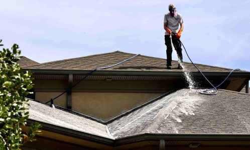 Person in protective gear pressure washes a sloped residential roof, spraying water onto dirty shingles under clear sky.